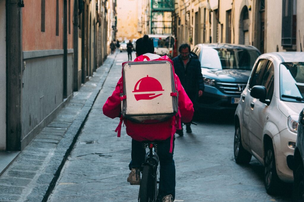 ヨーロッパ風の街中で配達バッグを背負って自転車を走らせている人の様子